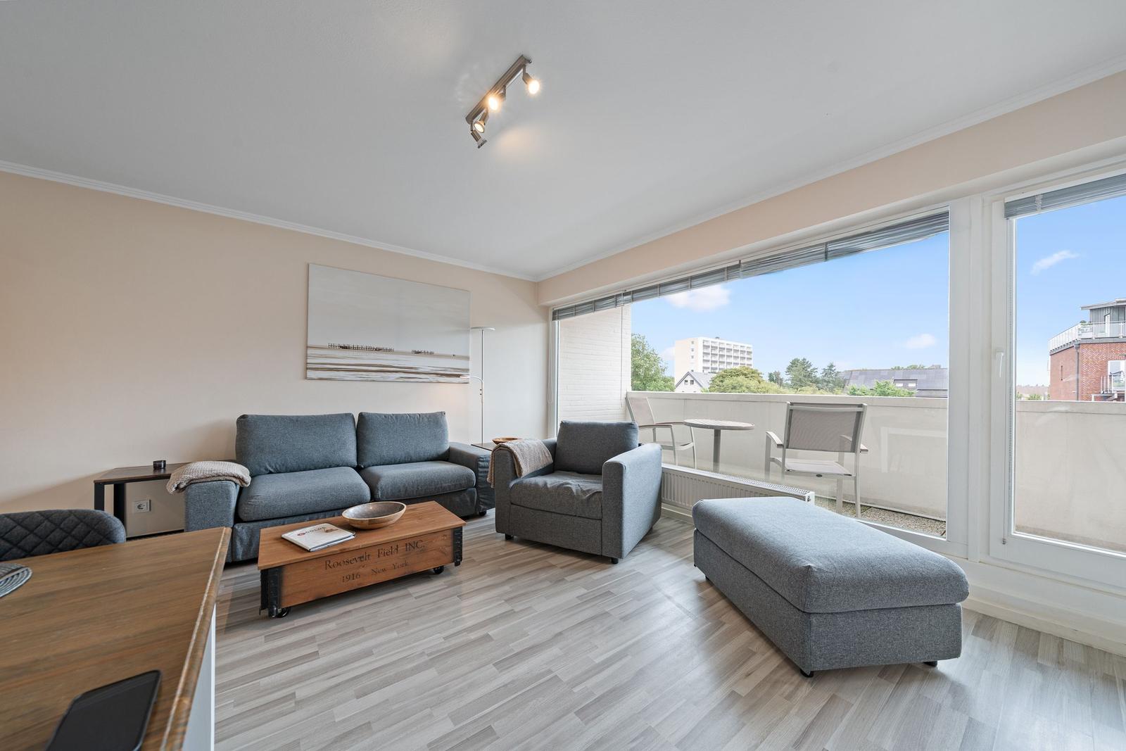 Wohnbereich mit Sofa, Sessel und Fußbank. Balkon mit Tisch und Stühlen. Holzboden und große Fenster.