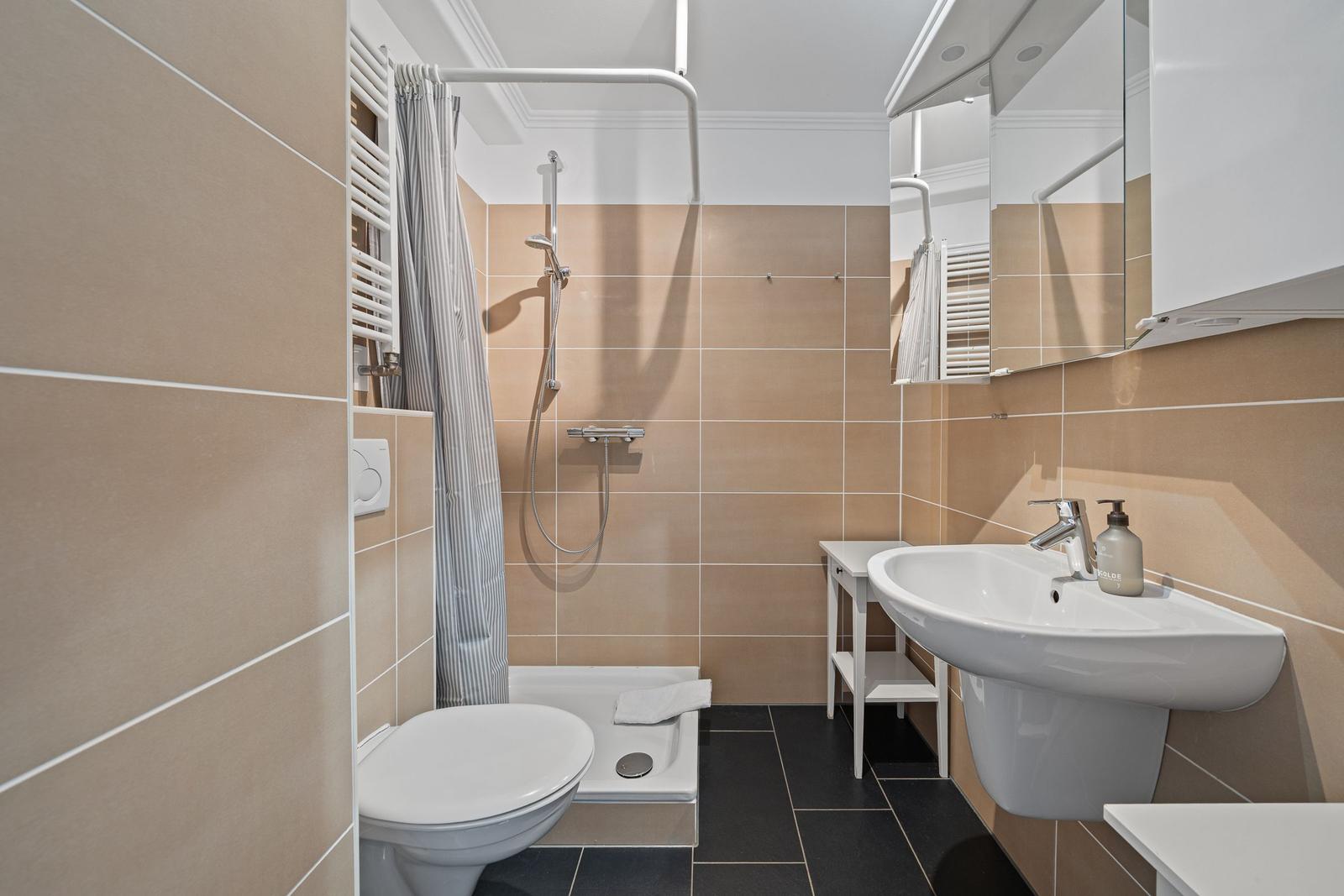 Badezimmer mit Dusche, Waschbecken und Toilette. Fliesenwand und dunkler Boden.