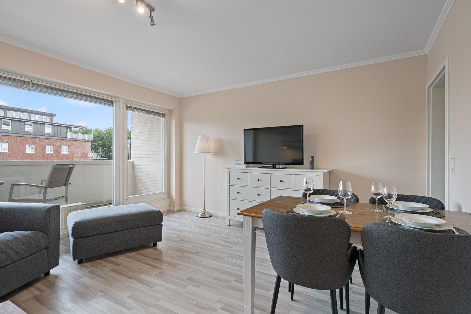Wohn- und Essbereich mit TV, Tisch und Stühlen sowie Balkon mit Sitzgelegenheit.