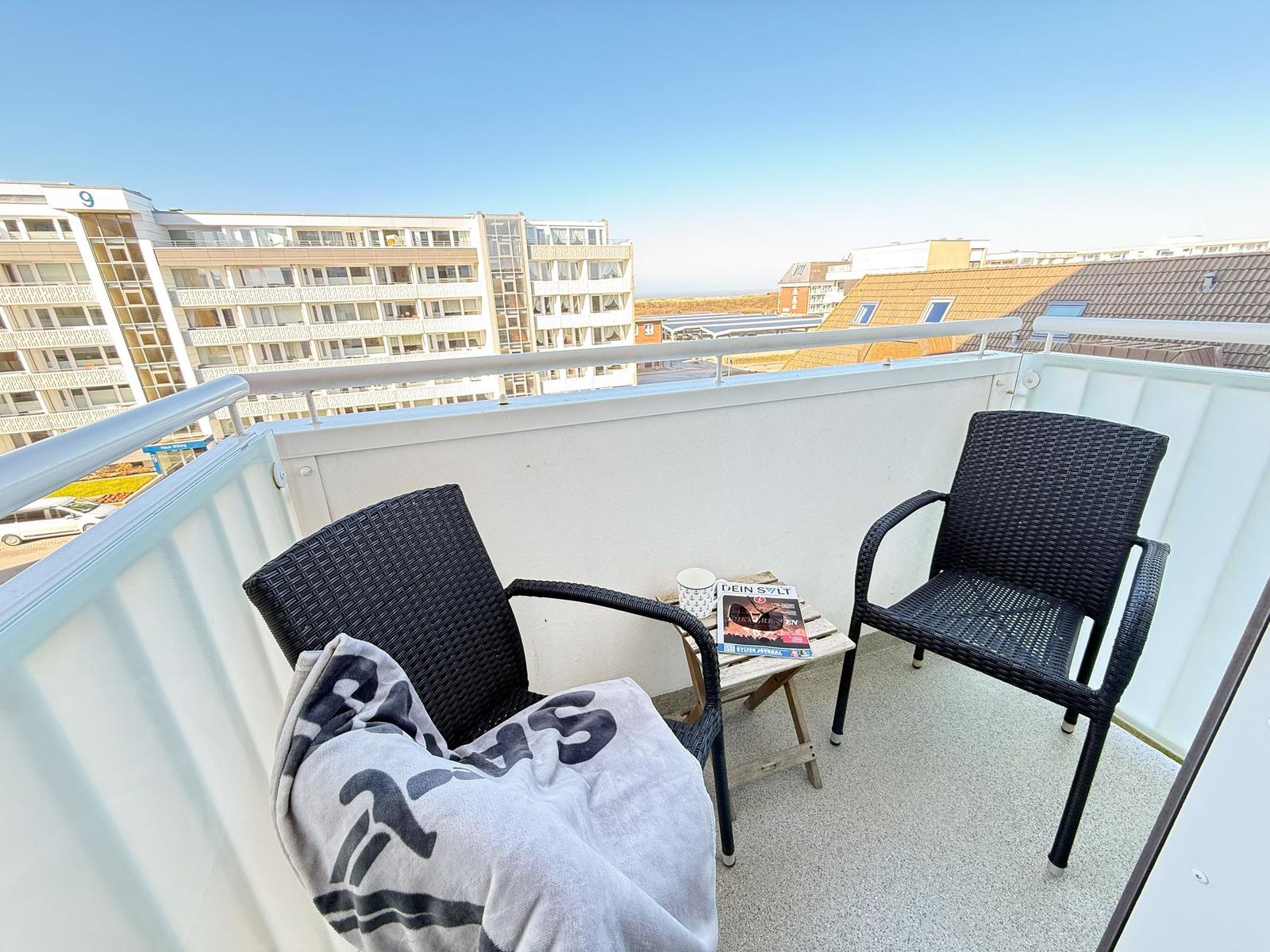 Balcony with two chairs, a small table, and a blanket. View of buildings and sky.