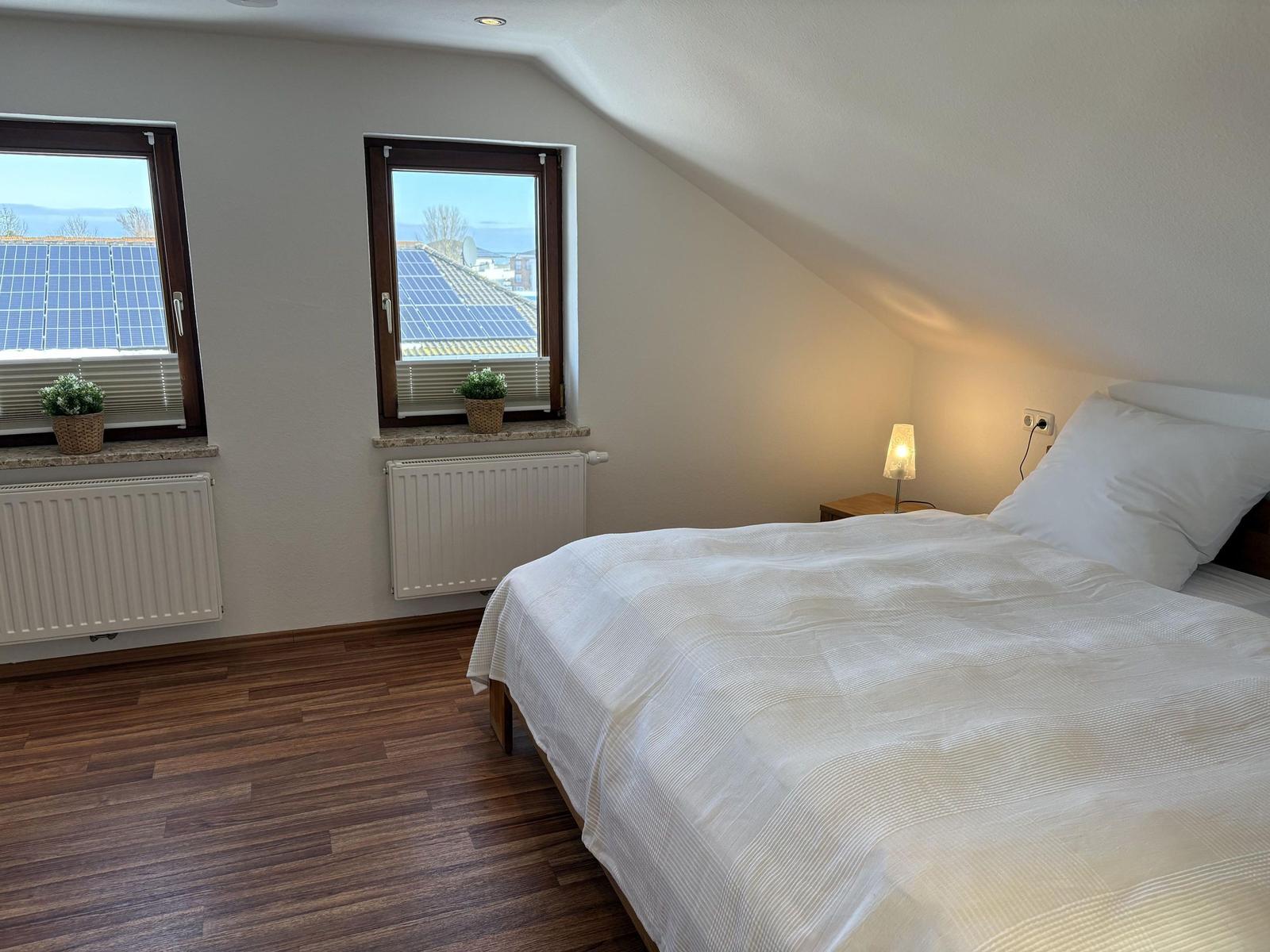 Schlafzimmer mit Dachschräge, zwei Fenstern und Holzboden. Bett mit weißer Bettwäsche.