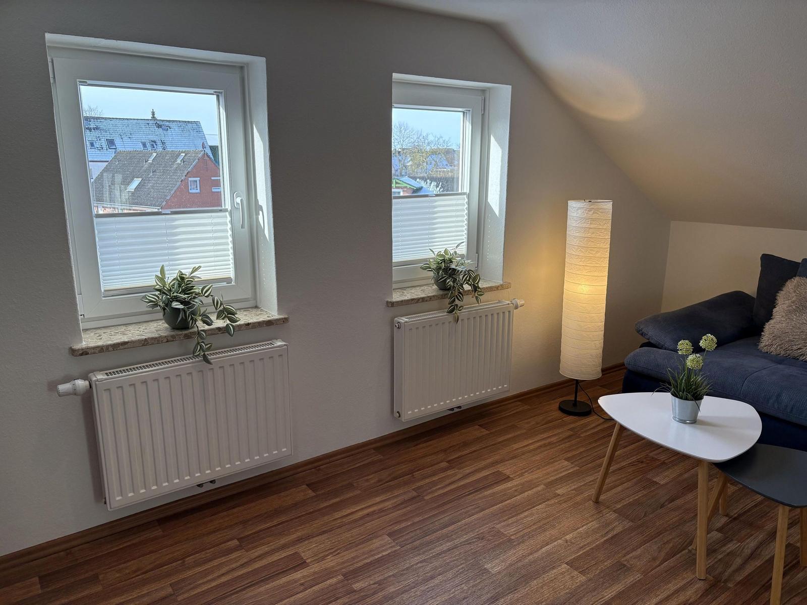 Wohnzimmer mit Sofa, Tisch, Lampe und zwei Fenstern mit Blick auf Dächer.