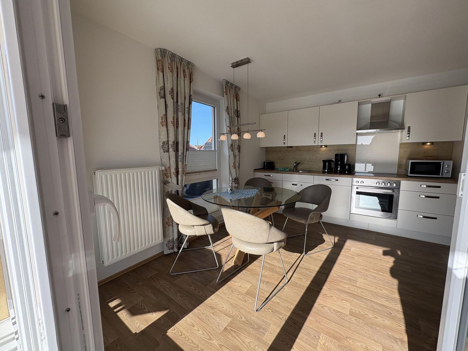 Küche mit Essbereich, Glas-tisch und vier Stühlen. Holzboden und Fenster mit Vorhängen.