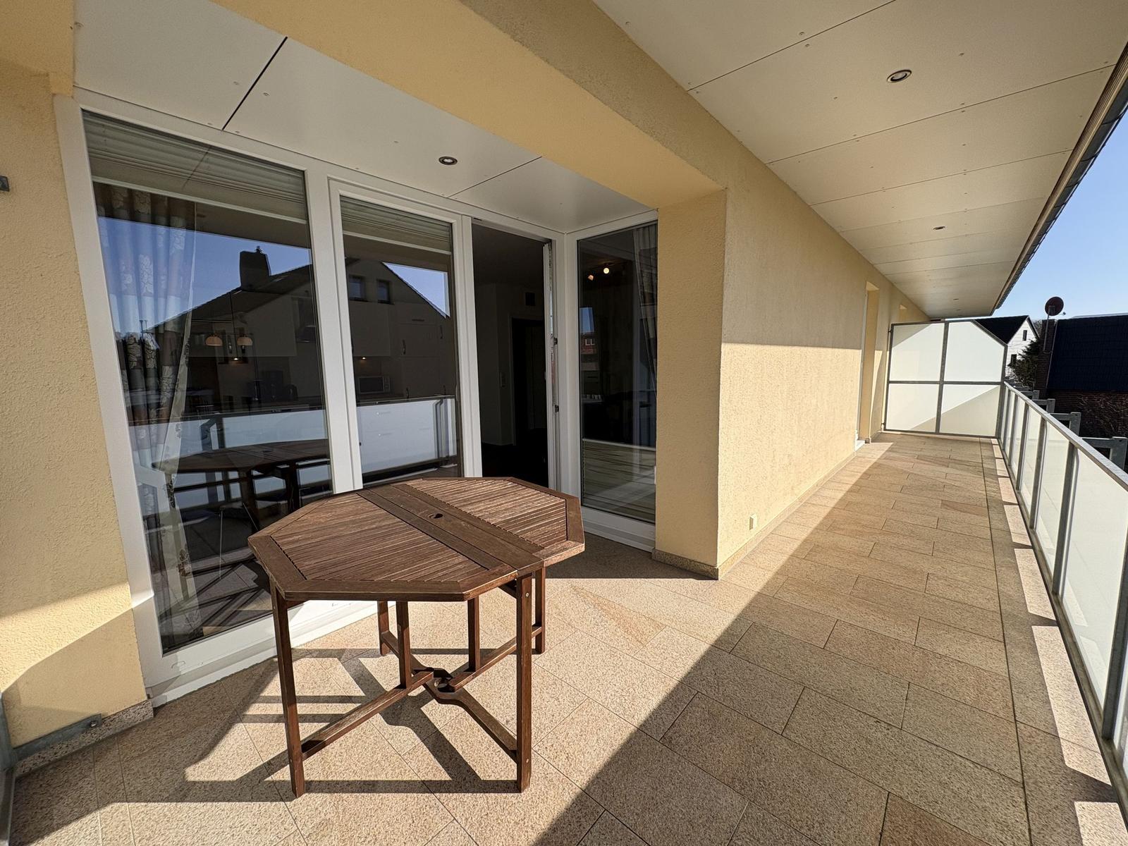 Terrasse mit Holztisch, Glasgeländer und Zugang ins Haus.