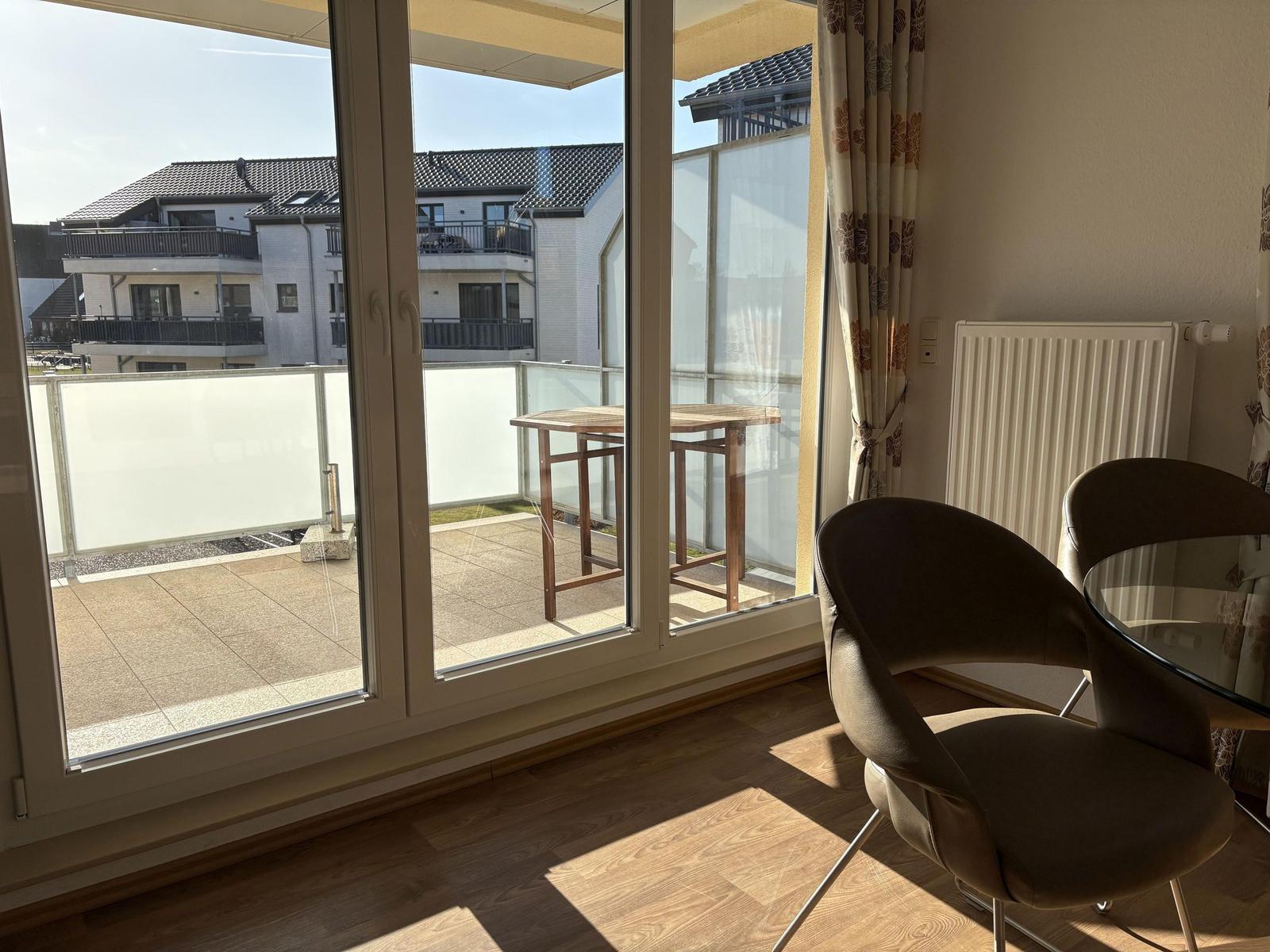 Durchgehendes Fenster mit Blick auf Balkon und Tisch. Holzboden und Stühle im Raum.