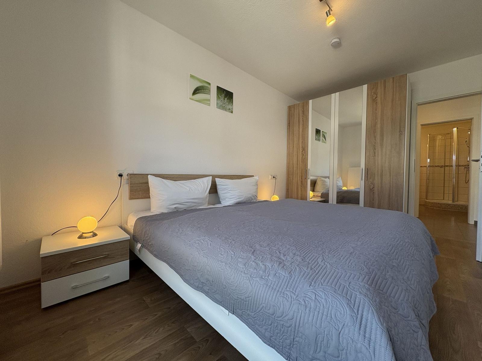 Schlafzimmer mit Doppelbett, Nachttisch und Kleiderschrank. Badezimmer mit Dusche ist sichtbar.