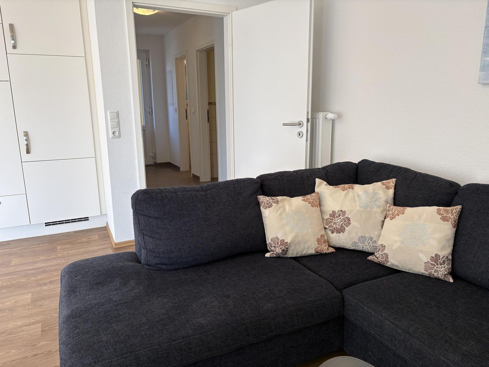 Schwarzes Sofa mit drei Kissen, Holzboden und Blick in den Flur.