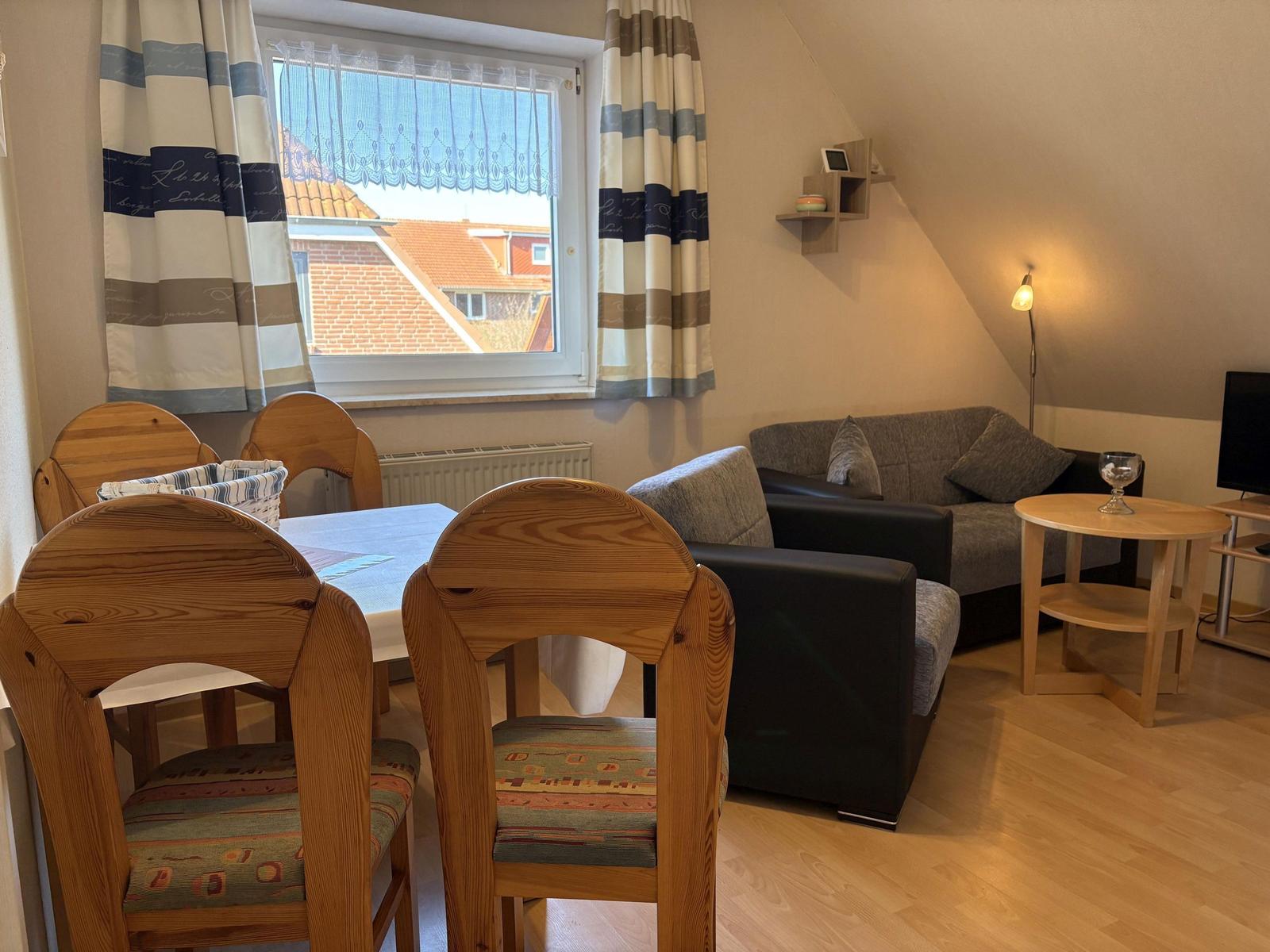 Wohn- und Essbereich mit Sofa, Tisch und Stühlen. Fenster mit Vorhängen und Blick auf Dächer.