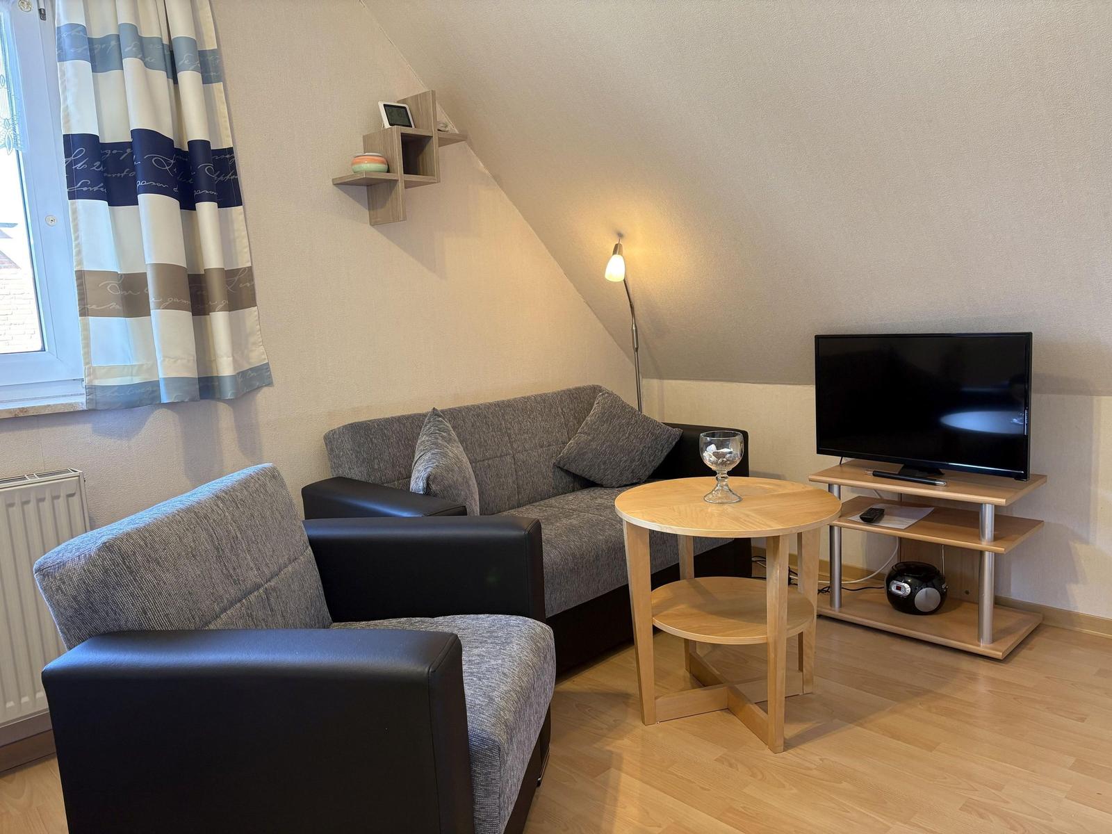 Wohnzimmer mit Sofa, Sessel, Tisch und Fernseher unter schräger Decke.