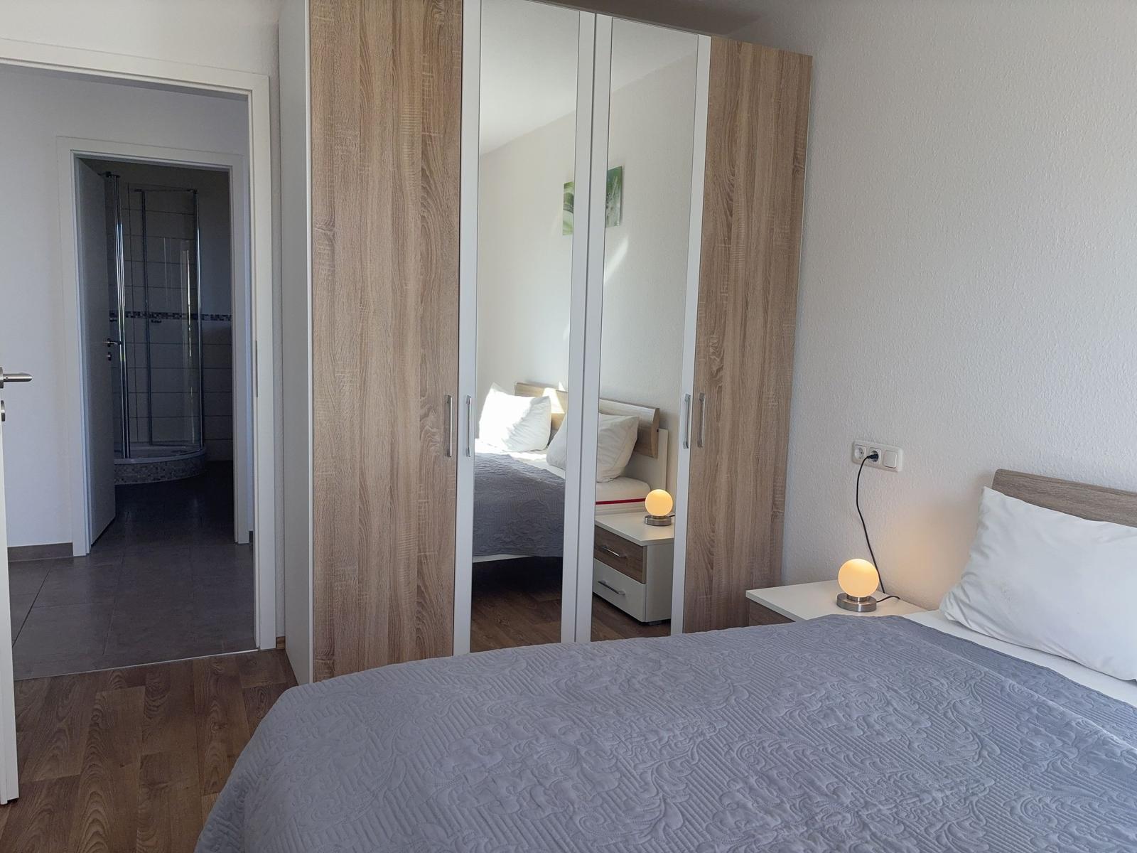 Schlafzimmer mit Doppelbett, Spiegelschrank und Nachttisch mit Lampe. Dusche im angrenzenden Raum.