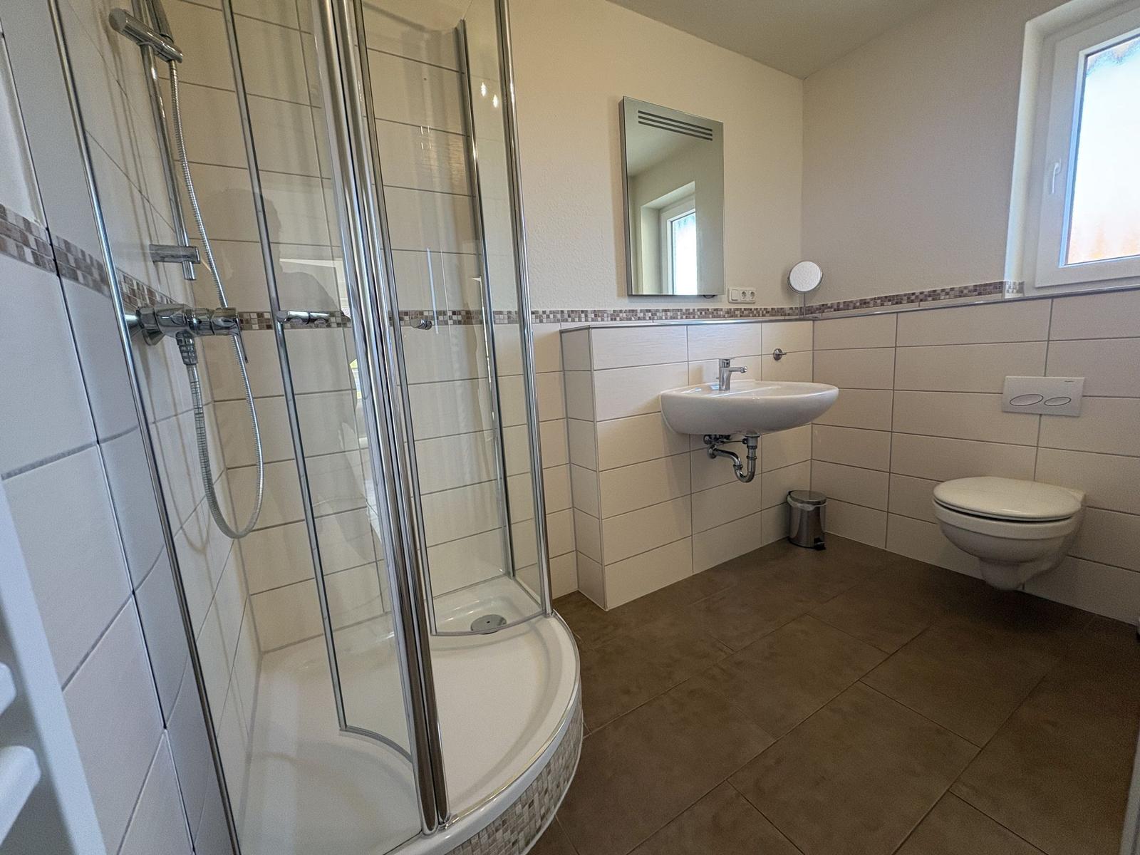 Badezimmer mit Dusche, Waschbecken und Toilette. Fliesenwand und Fenster.