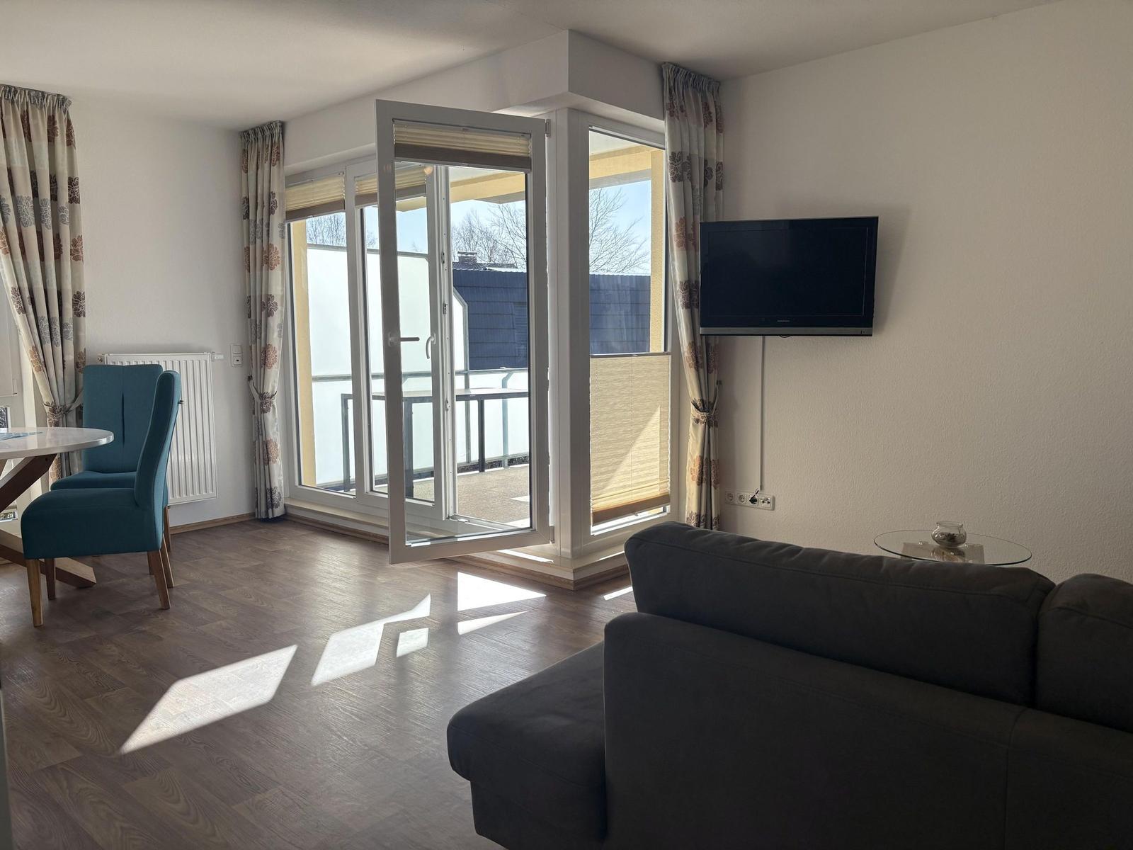 Wohnraum mit Sofa, TV und Balkon. Holzboden und Fenster mit Vorhängen.