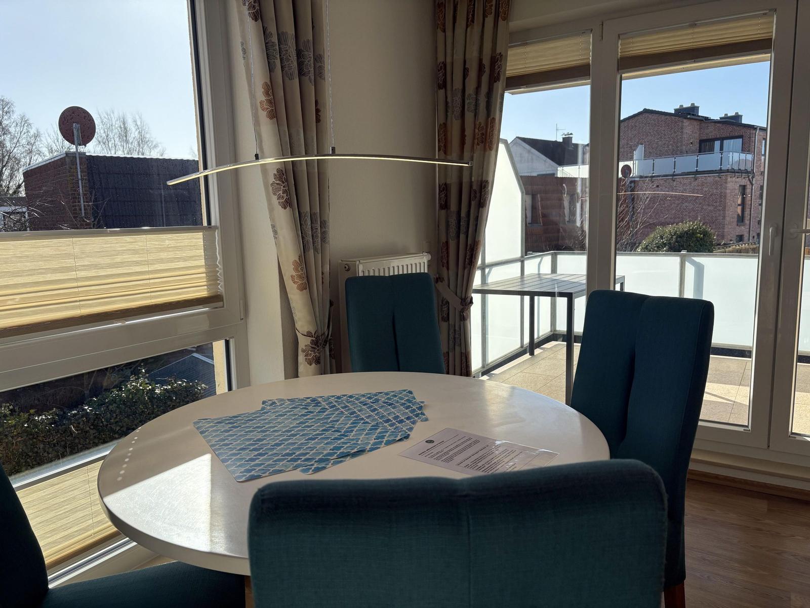 Runder Tisch mit Stühlen und Blick auf Balkon. Vorhänge und Fenster sind sichtbar.