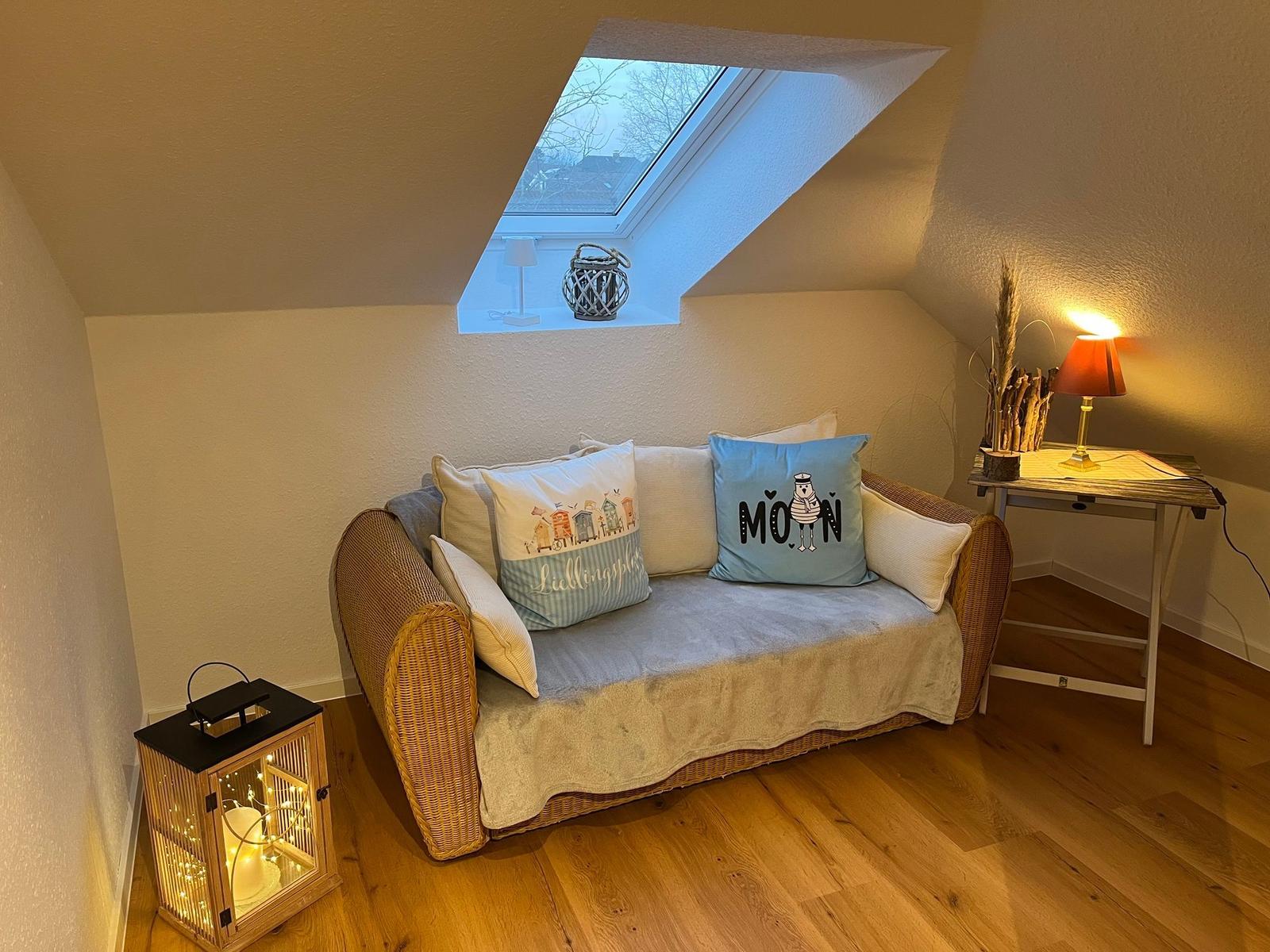 Ein Sofa mit Kissen unter einem Dachfenster. Neben einem Tisch mit Lampe und einer Laterne.