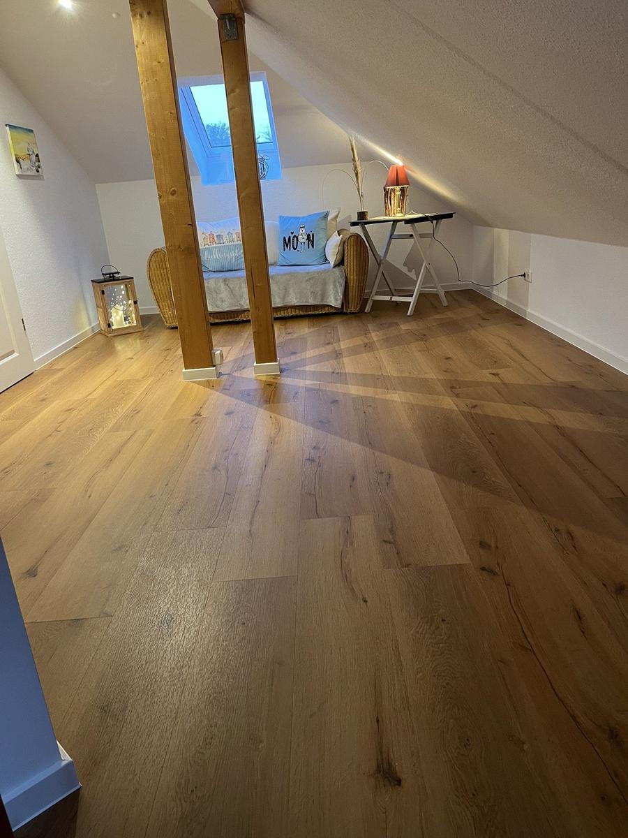 Schlafzimmer mit Holzboden, Dachschräge und Fenster. Möbel und Beleuchtung sind sichtbar.
