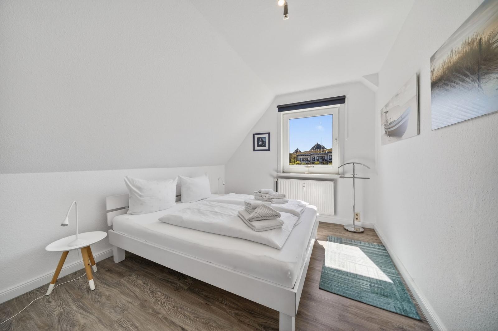 Schlafzimmer mit weißem Bett, Holzboden und Fenster mit Blick nach draußen.
