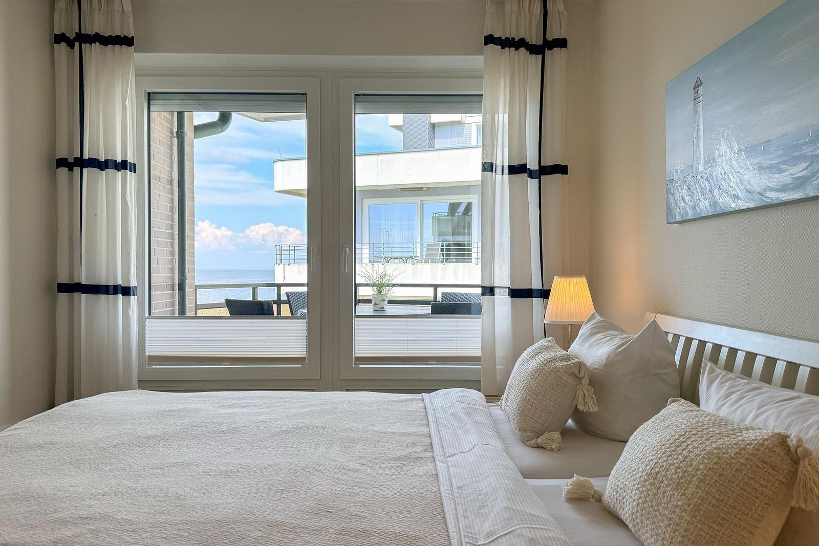 Schlafzimmer mit Blick auf Balkon und Meer. Bett, Lampen und Wandbild mit Leuchtturm.