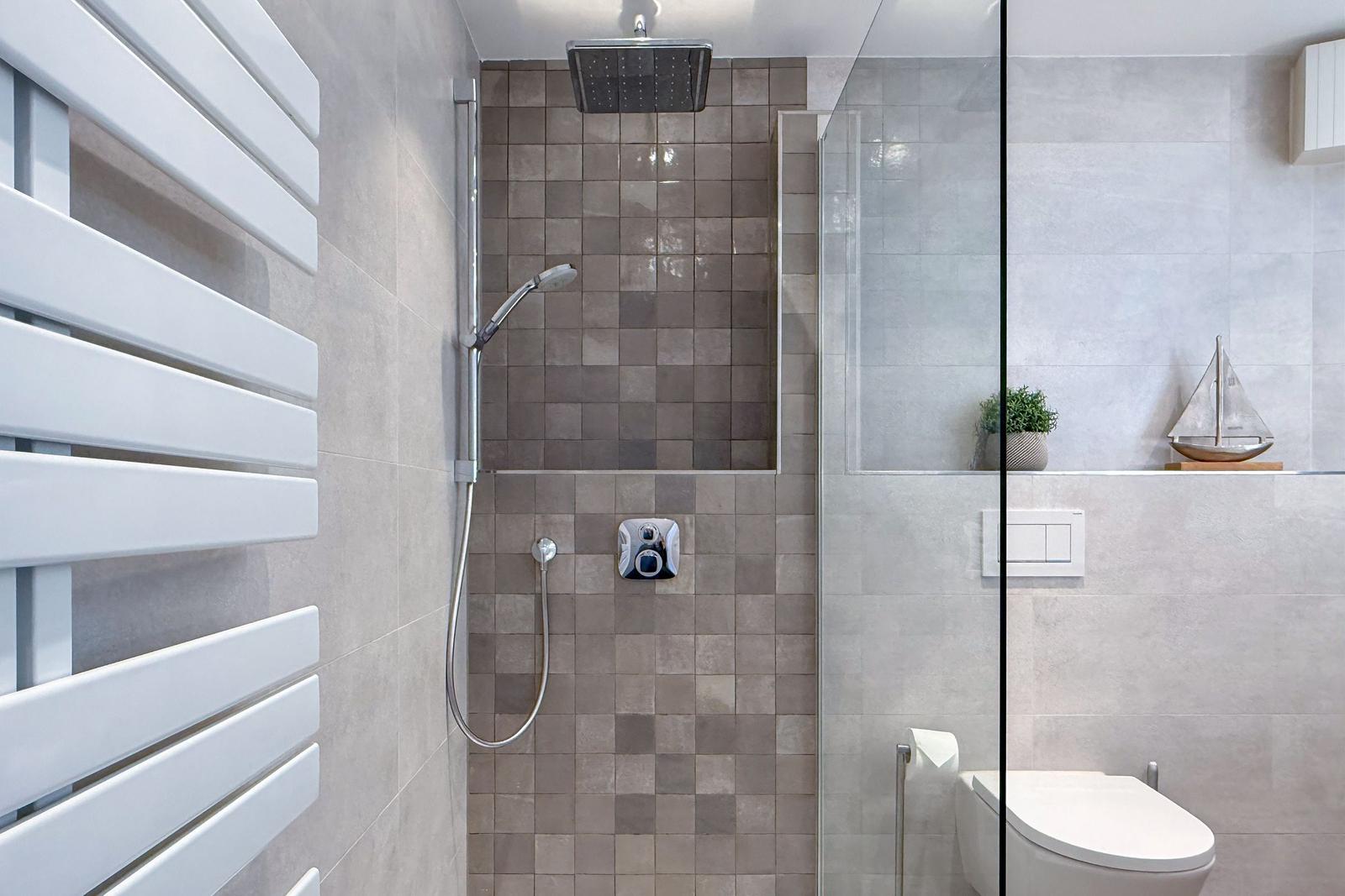 Dusche mit Duschbrause, Toilette und Heizung. Wandfliesen in Grau.