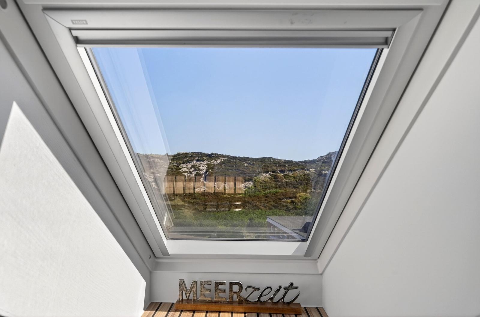 Dachfenster mit Blick auf Hügel und Himmel. Unten steht MEERzeit auf Holz.