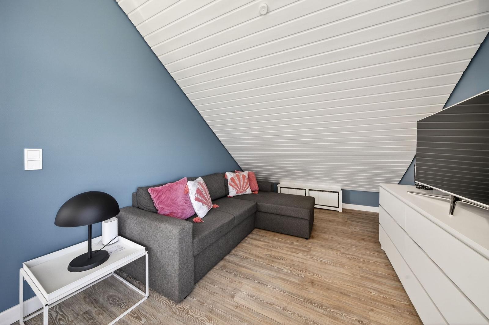 Wohnbereich mit grauem Sofa, rosa Kissen, Holzboden und weißer Schrankwand.