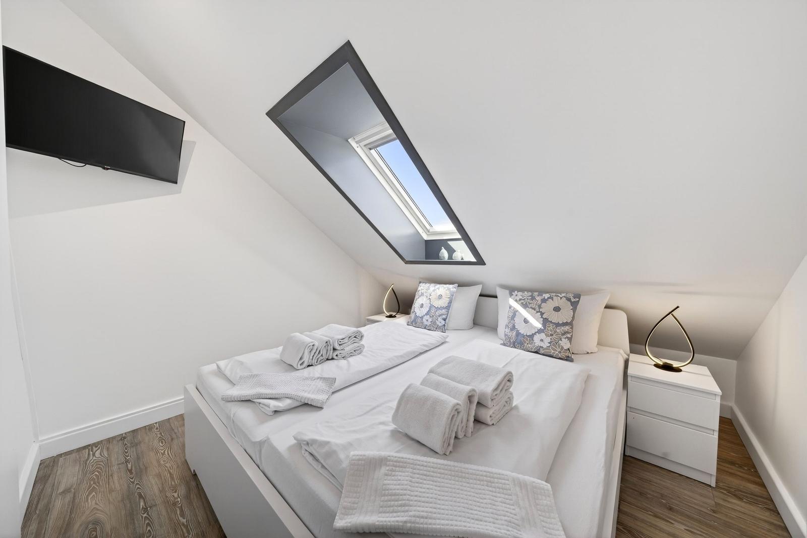 Schlafzimmer mit Dachfenster, Bett, Nachttisch und Fernseher an der Wand.
