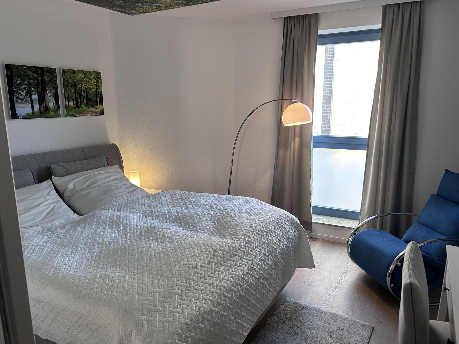 Schlafzimmer mit Bett, Stehlampe und blauem Sessel. Fenster mit Vorhängen.
