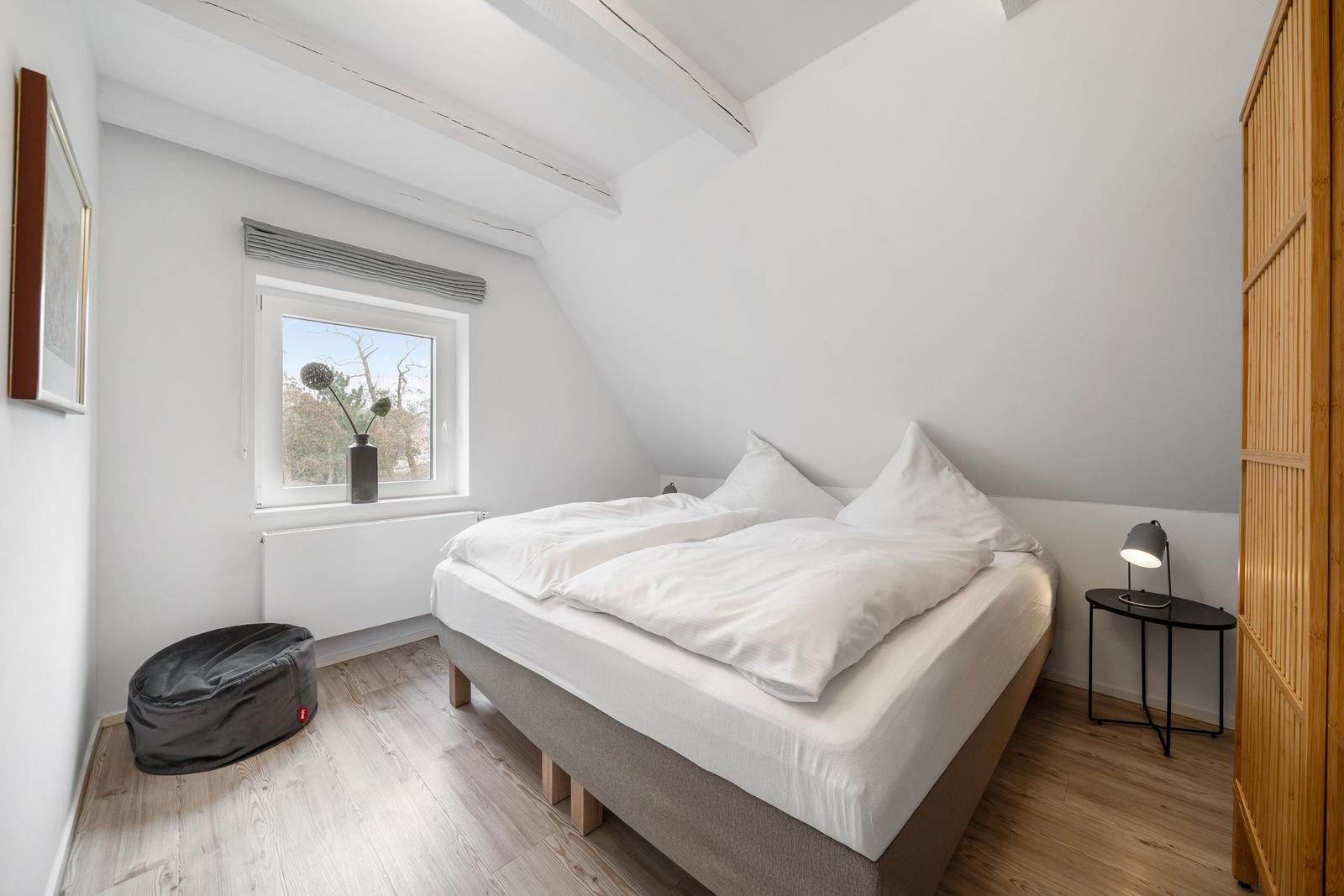 Bedroom with double bed, window, and wooden floor. Beside the bed is a nightstand with lamp.
