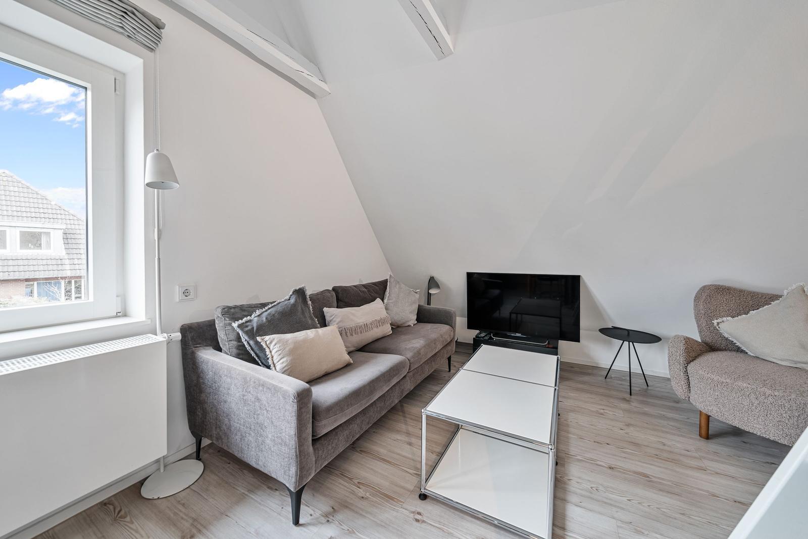 Living room with sofa, armchair, TV, and window. Wooden floor and sloped ceiling.