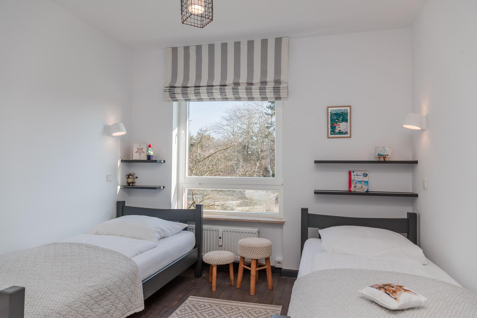 Two single beds with white linens and blankets. Window with striped roller blind. Wooden stools and wall shelves.