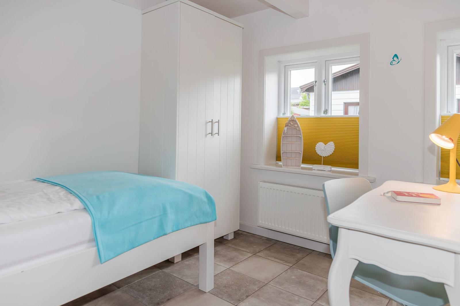 Schlafzimmer mit weißem Bett, Schrank und Schreibtisch. Fenster mit gelbem Jalousie.