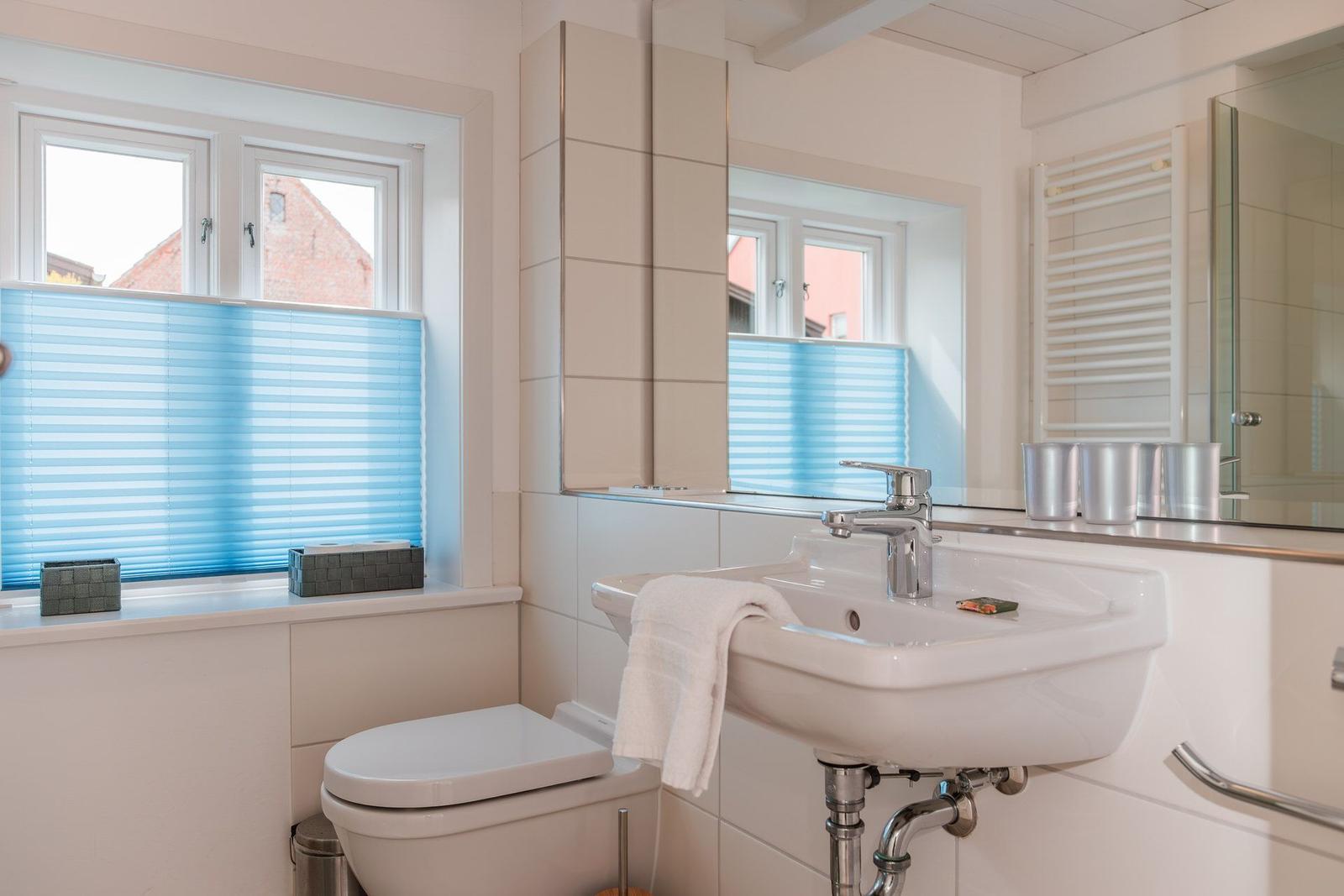 Badezimmer mit Waschbecken, Spiegel, Toilette und Fenster mit blauen Jalousien.