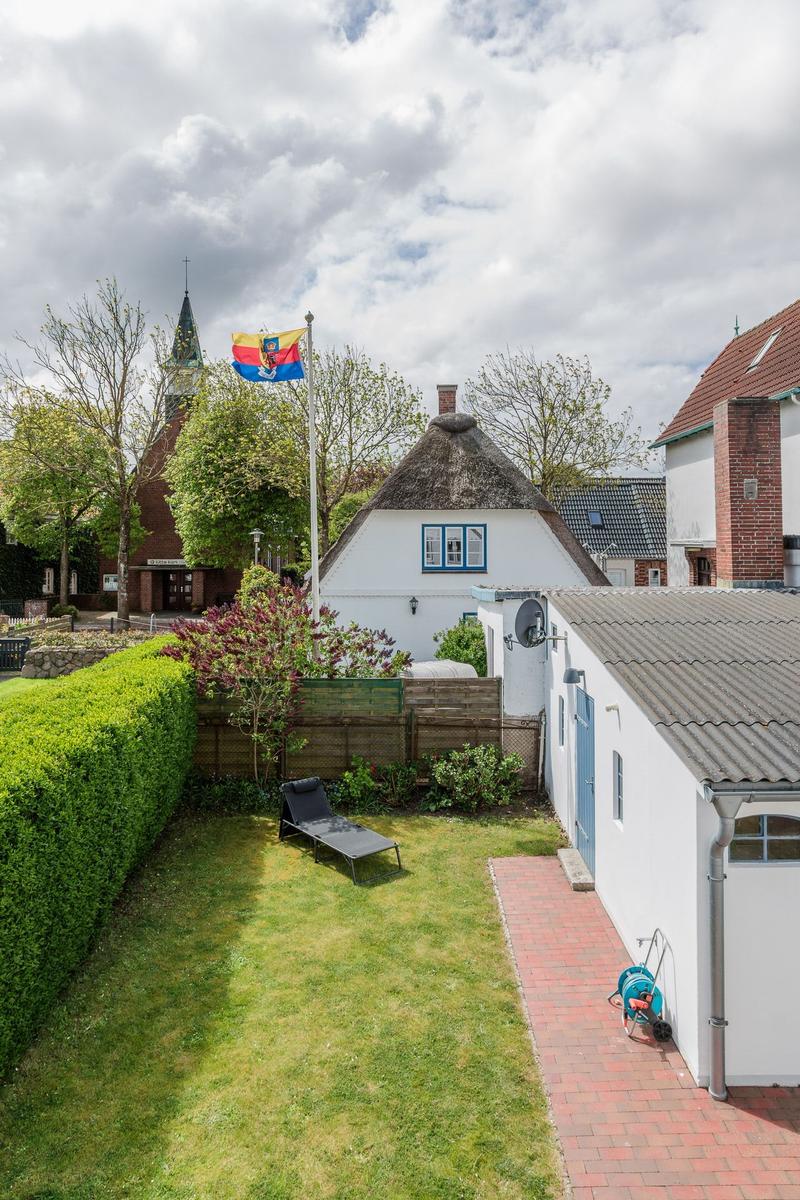Hinterhof mit Garten, Liegestuhl und Flagge vor einem weißen Haus mit Strohdach.