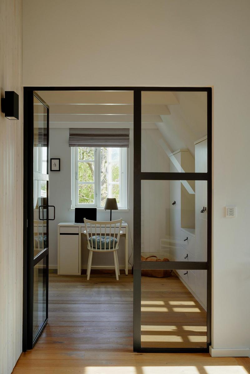 Durchgehende Glasschiebetüren führen in ein Arbeitszimmer mit Schreibtisch und Fenster.