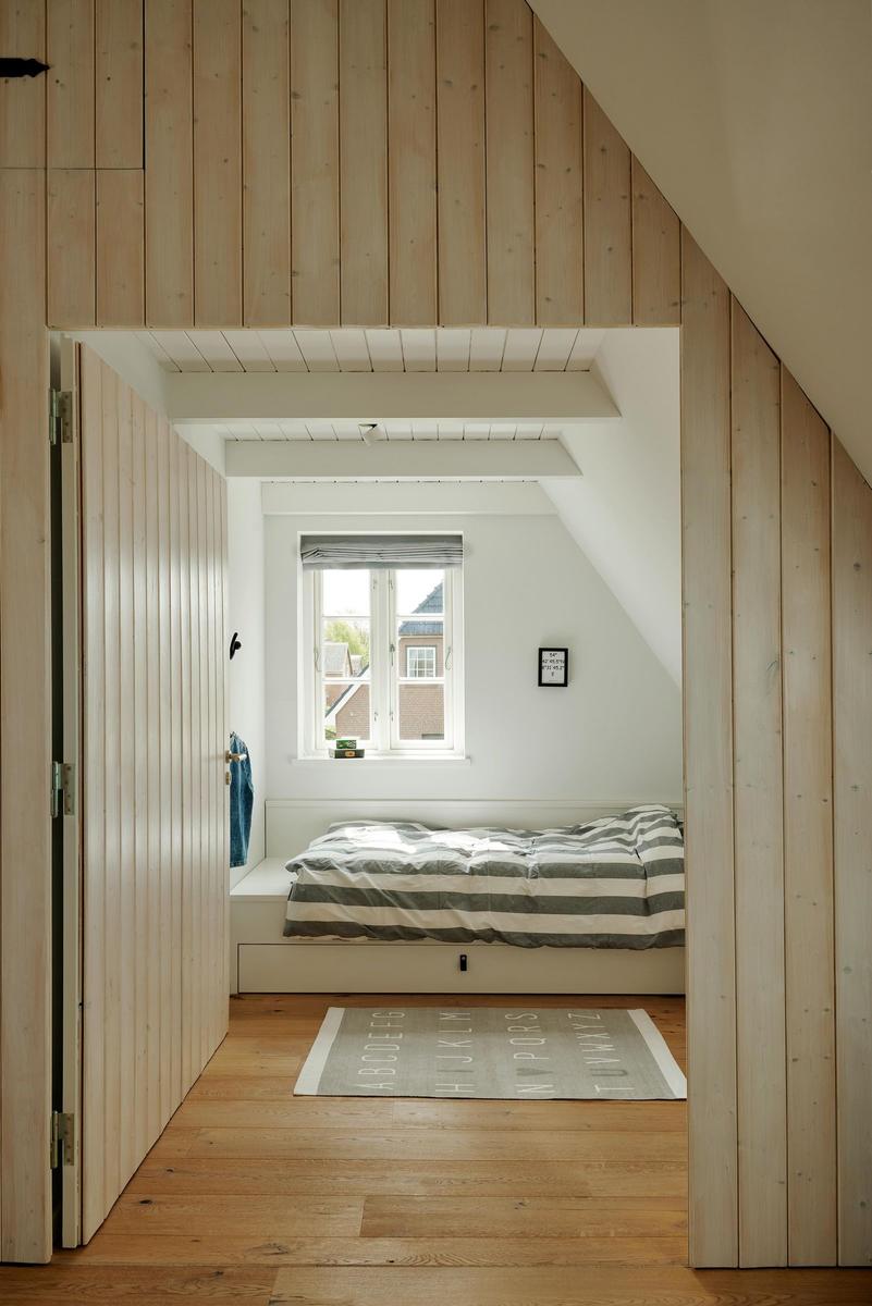 Schlafzimmer mit Dachschrägen, Holzwand und Fenster. Bett mit Streifenbezug und Teppich mit Buchstaben.