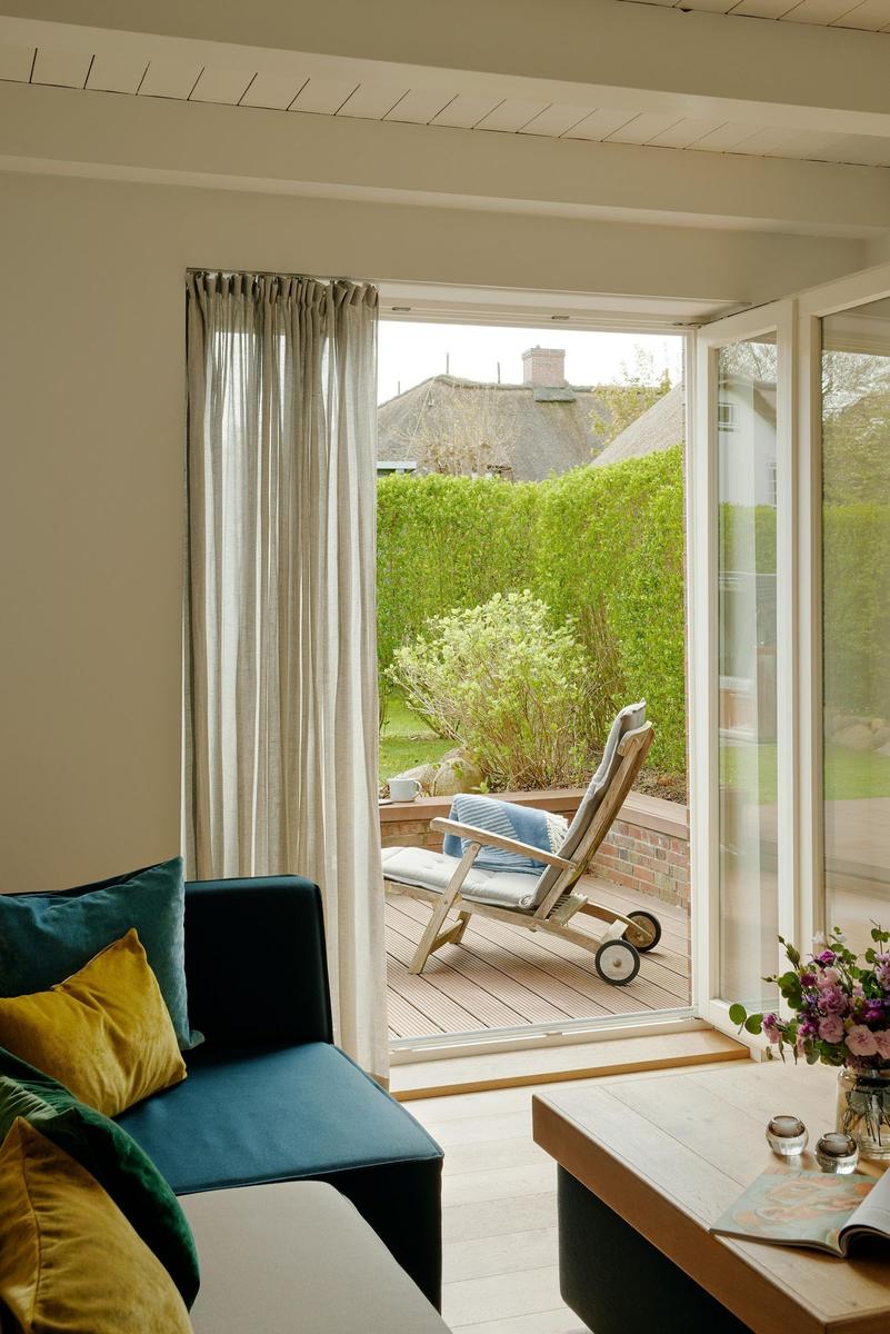 Ein Wohnbereich mit Blick auf eine Terrasse und einen Garten. Ein Sofa mit Kissen und ein Tisch sind sichtbar.