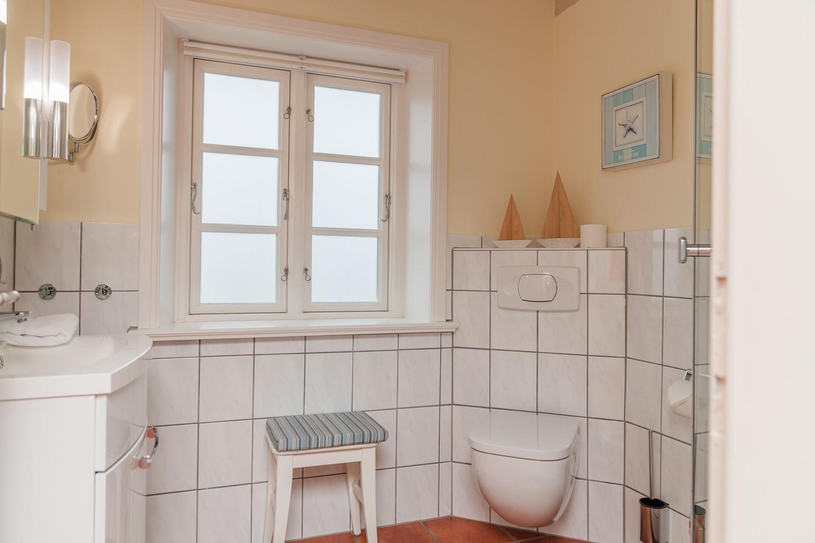 Badezimmer mit Toilette, Waschbecken und Fenster. Wandfliesen und Holzstuhl.