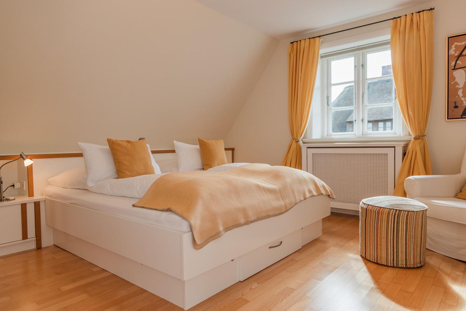 Schlafzimmer mit Doppelbett, gelben Vorhängen und Holzboden. Fenster zeigt Dachhäuser.