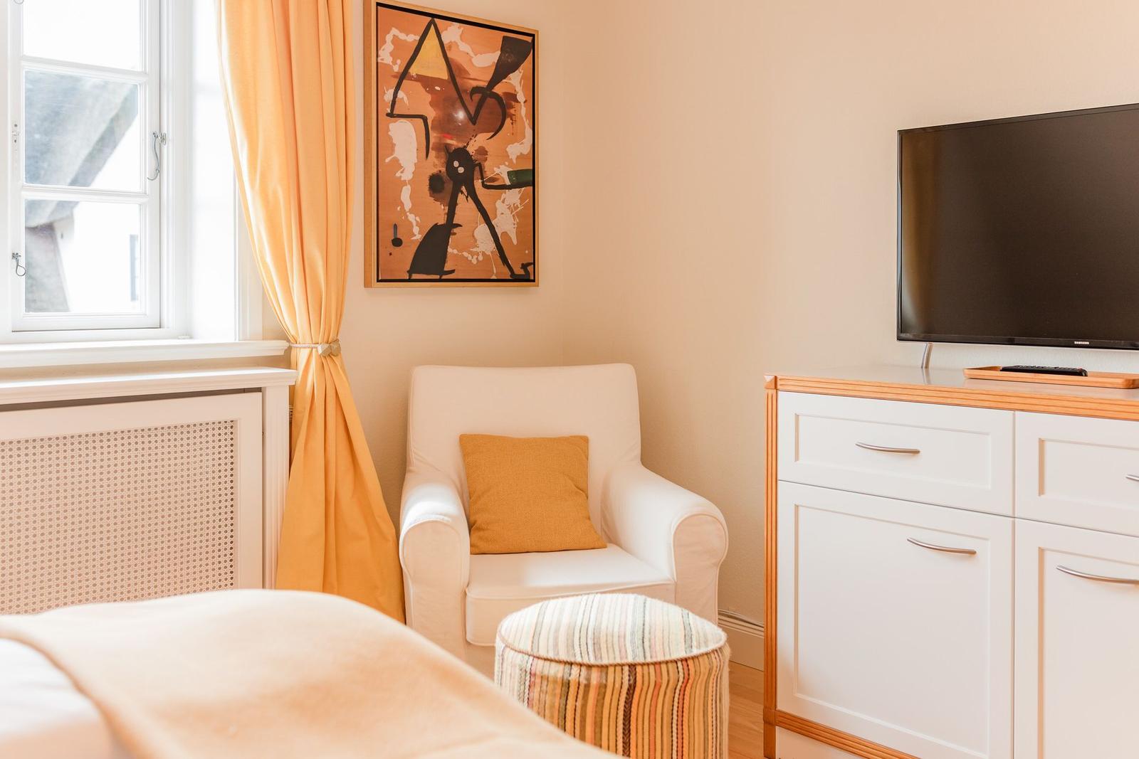 Ein Zimmer mit weißem Sessel, orangefarbenen Vorhängen und Fernseher auf Schrank.