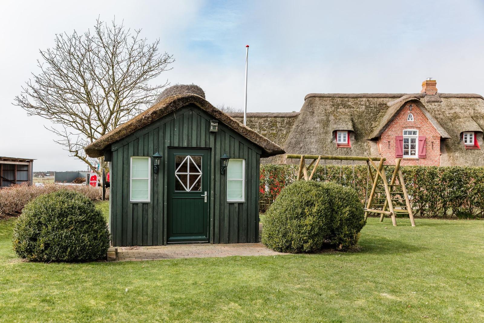 Ein grünes Holzhaus mit Strohdach und eine größere Villa mit Strohdach im Hintergrund.