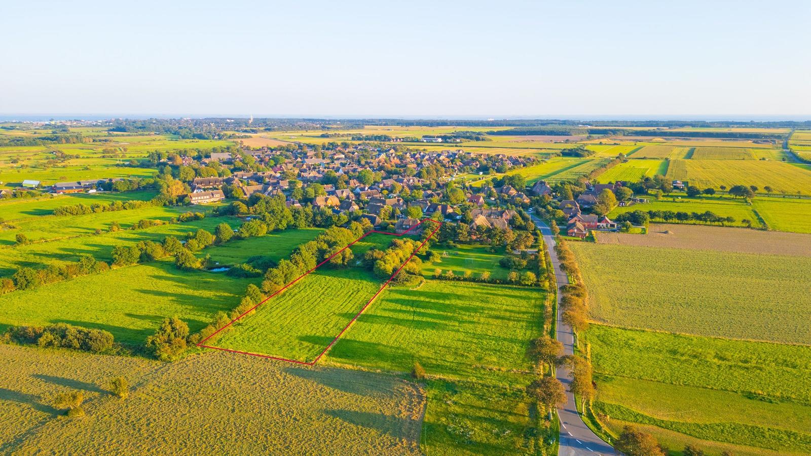 Grünes Feld mit roter Linie, Dorf im Hintergrund, Straße und Bäume
