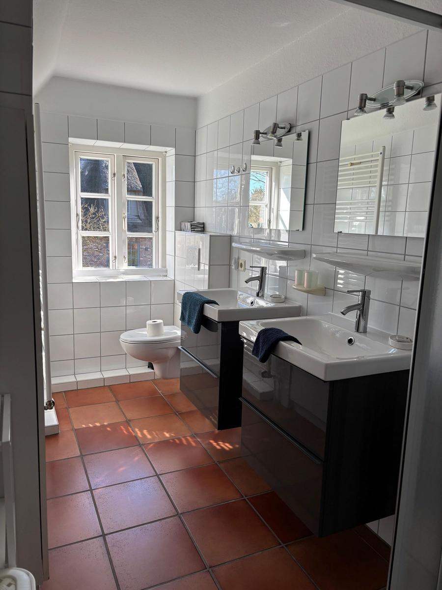 Badezimmer mit zwei Waschbecken, Toilette und Fenster. Fliesenboden und Wandfliesen.
