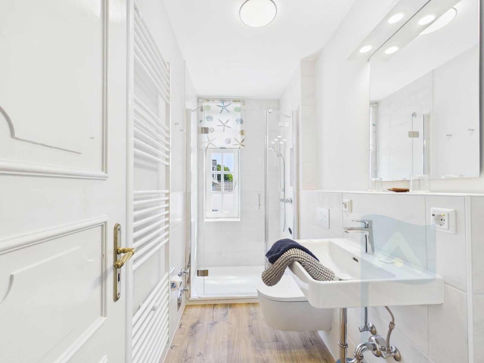 Badezimmer mit Dusche, Waschbecken und Spiegel. Holzboden und Fenster mit Seestern-Motiv.