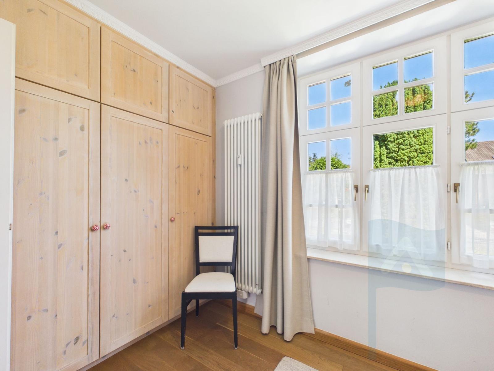 Ein Zimmer mit Holzschrank, Stuhl und Fenster mit Blick auf Grün.