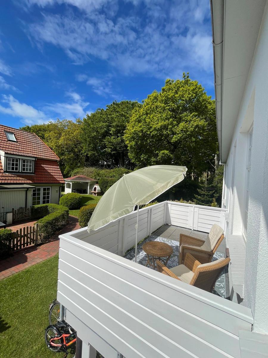 Terrasse mit Sonnenschirm, zwei Stühlen und Tisch. Blick auf grüne Bäume und ein Haus.