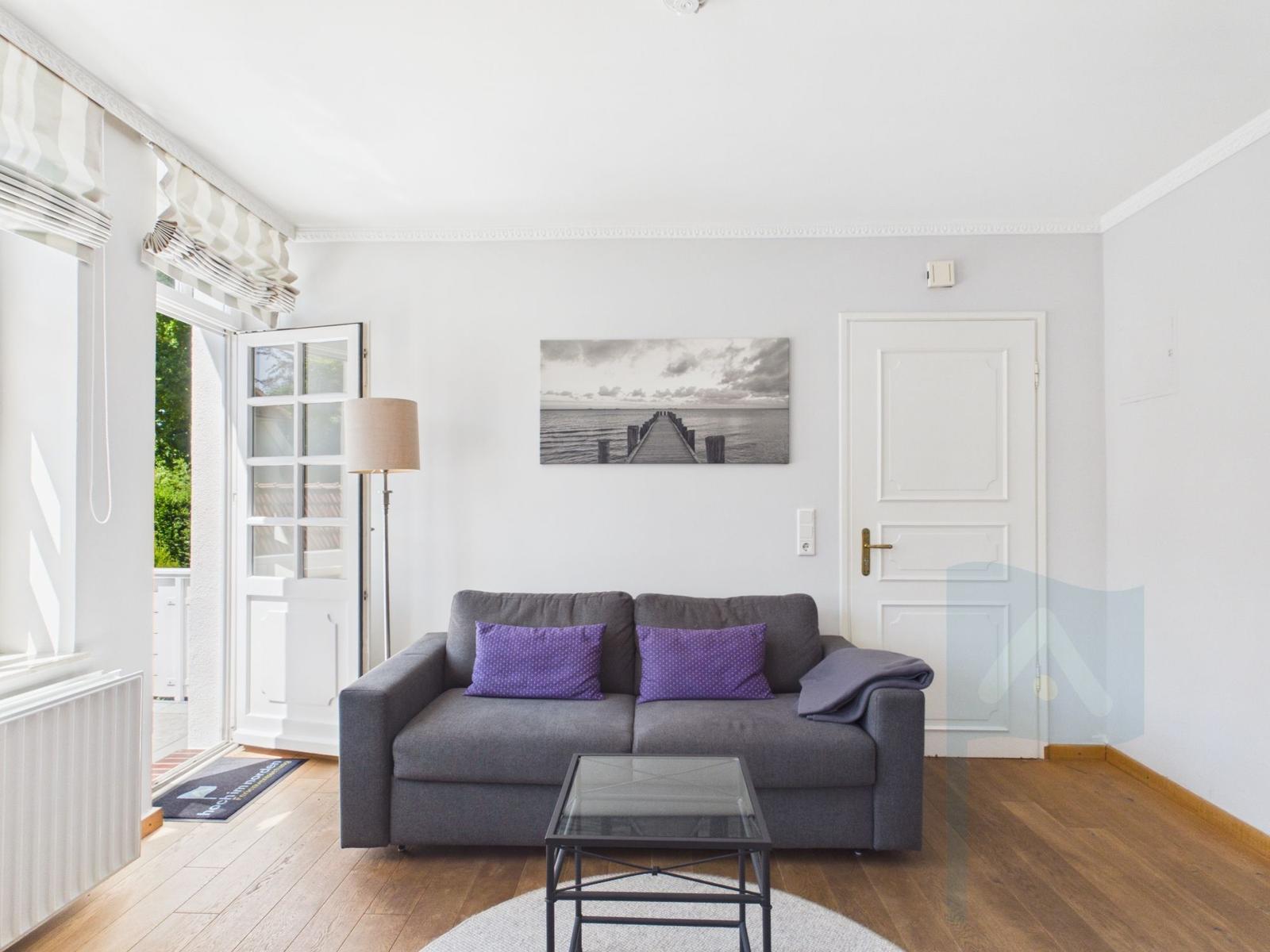 Ein Wohnzimmer mit grauem Sofa, zwei lila Kissen und einem Glas-Tisch.