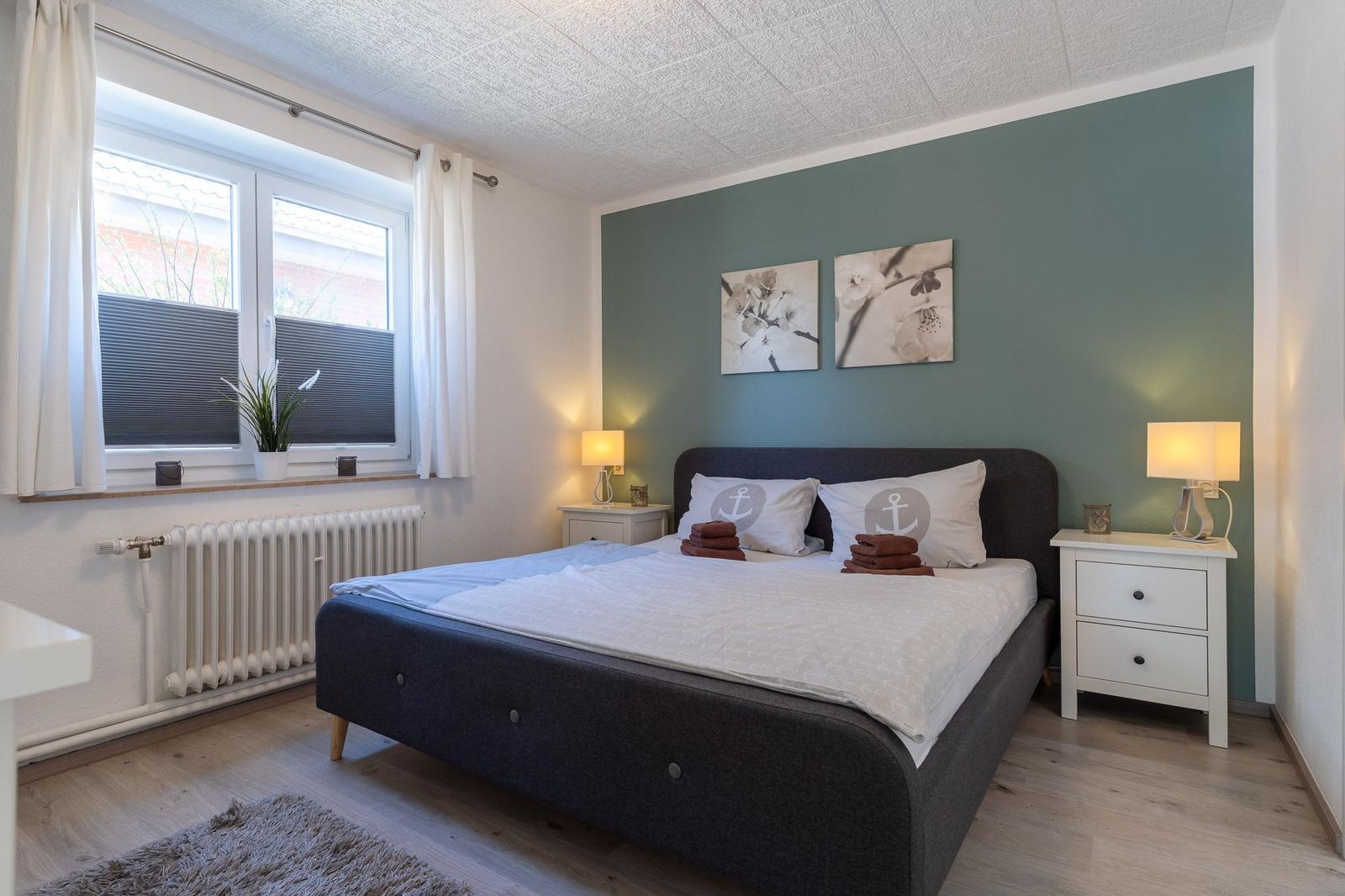 Schlafzimmer mit Bett, zwei Nachttischen und Fenster. Wand in Grün mit Bildern.