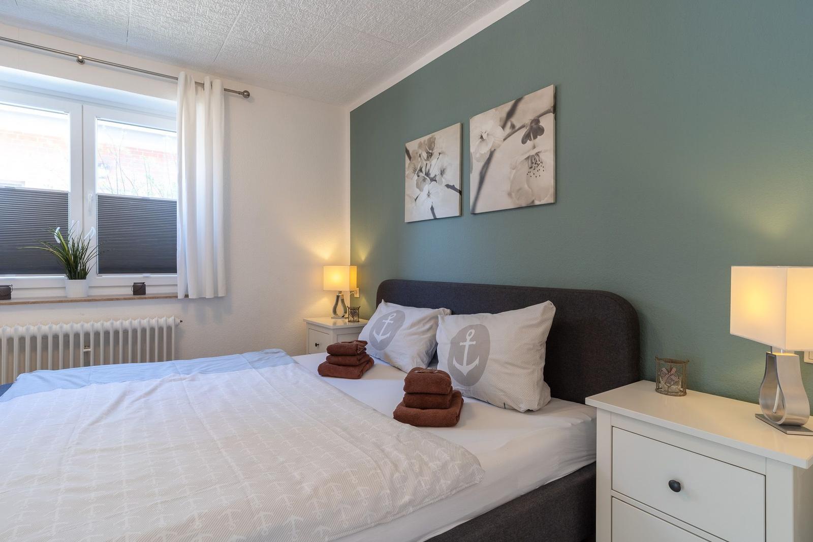 Schlafzimmer mit Bett, Nachttischen und Fenster. Wand mit Bildern. Bett mit Kissen und Handtüchern.