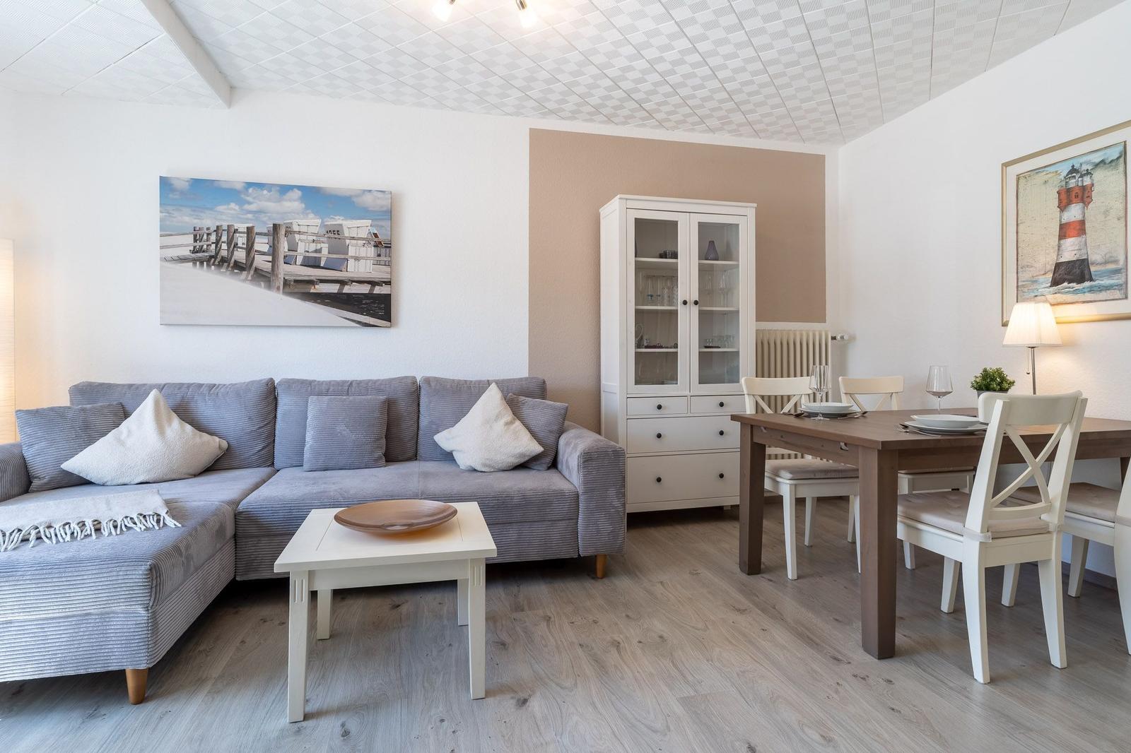 Wohn- und Essbereich mit Sofa, Tisch und Schrank. Wandbilder zeigen Leuchtturm und Strand.
