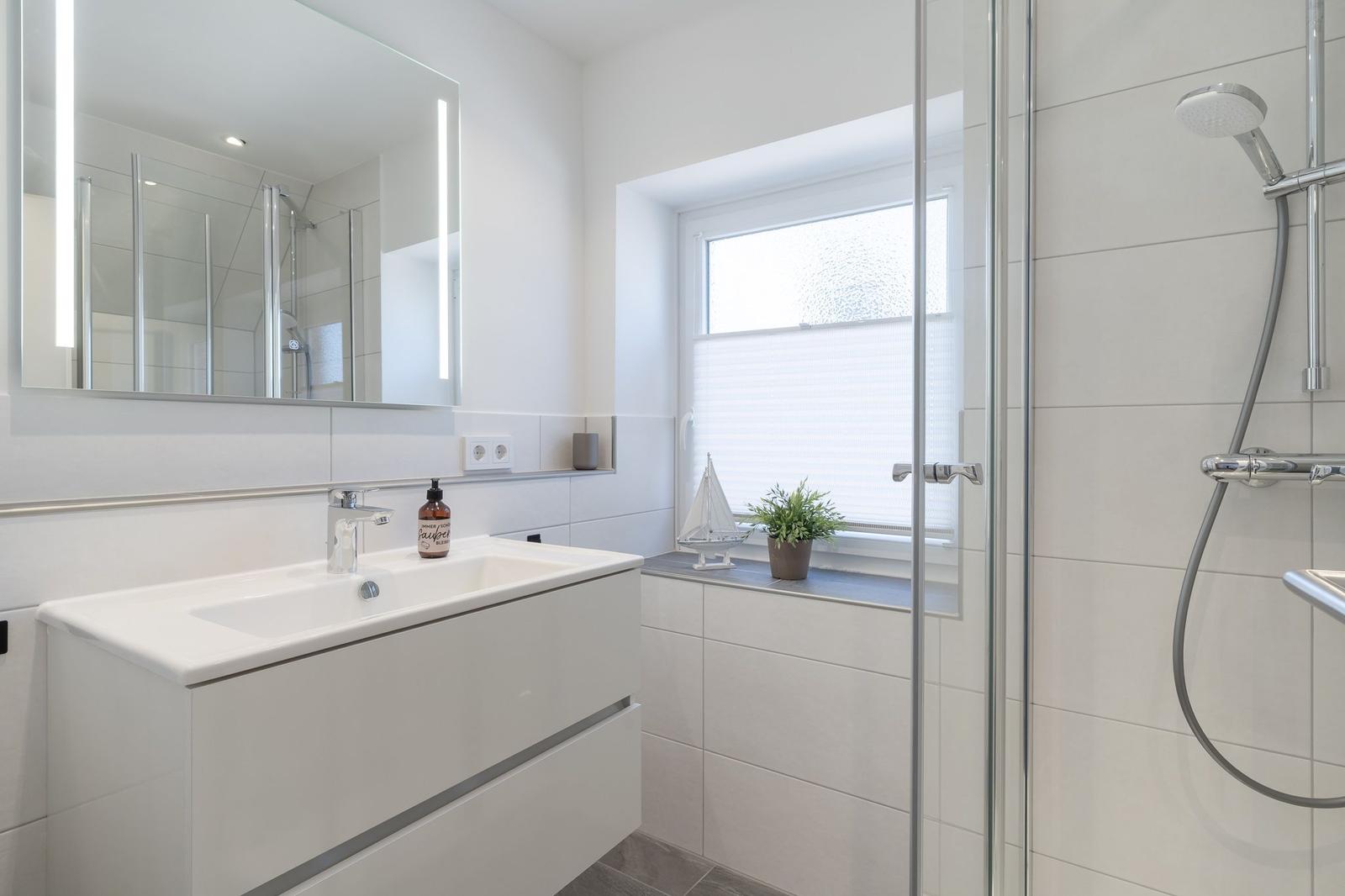 Badezimmer mit Dusche, Waschbecken und Spiegel. Fenster mit Pflanze und Segelboot.