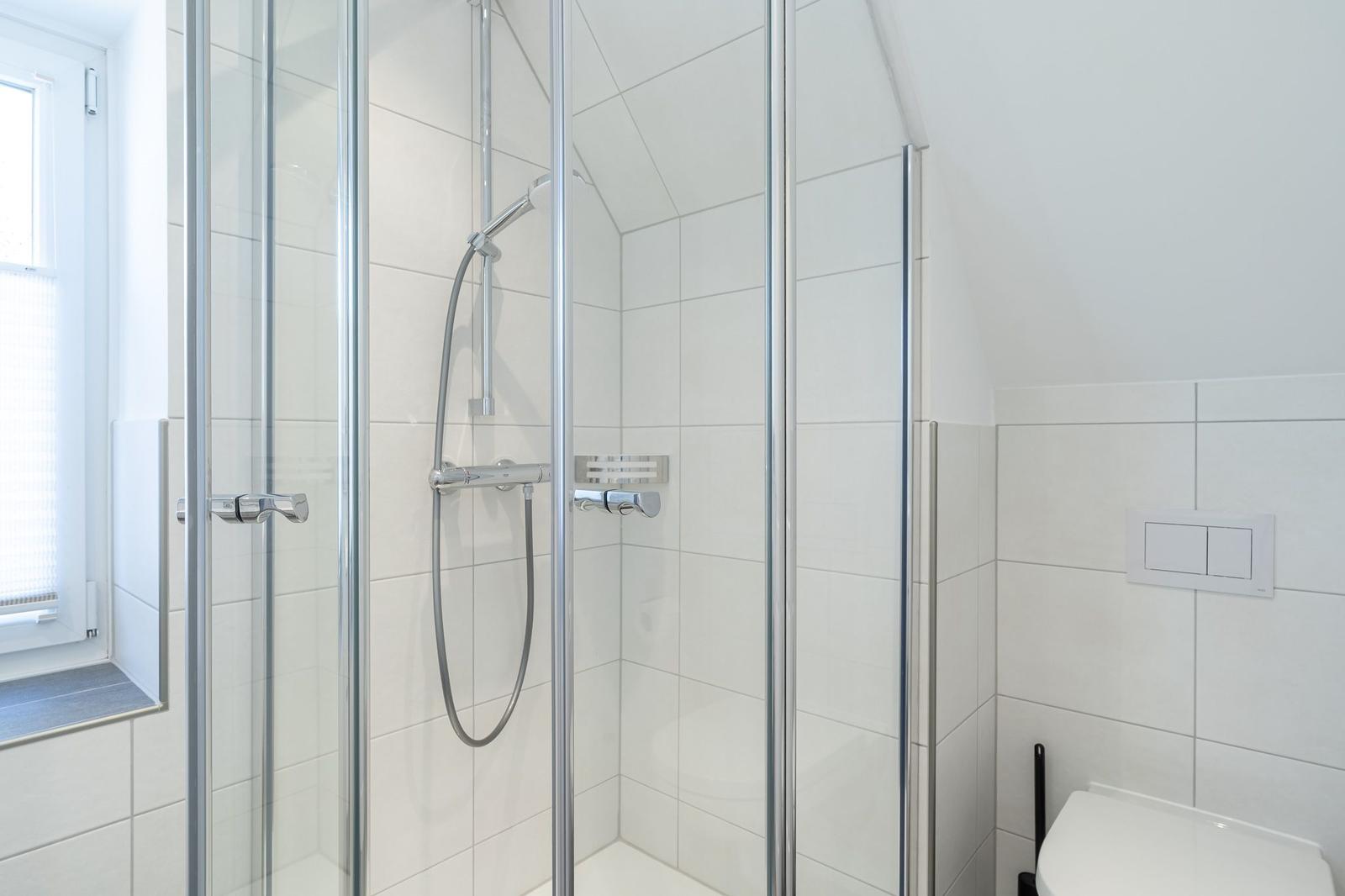 Dusche mit Glaswand, Toilette und Fenster in weißer Fliesenwand.